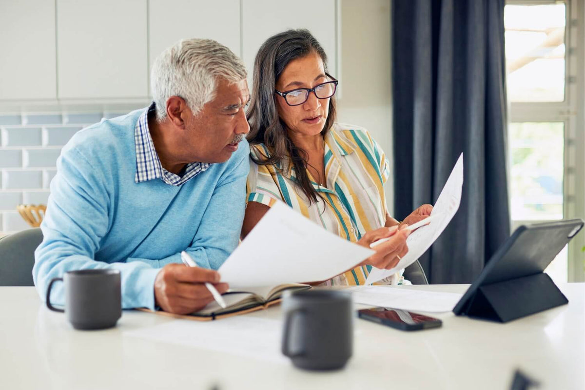Two middle-aged adults sit at a kitchen table reviewing paperwork together. One person holds a pen and papers while the other reads a document; a tablet, smartphone, notebooks, and coffee mugs are on the table, suggesting shared financial or household pla