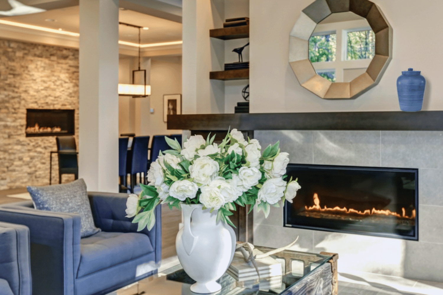 Modern living room with a cozy fireplace built into a tiled wall. Above the fireplace is a decorative, geometric-framed round mirror, and to the right sits a blue ceramic urn. In the foreground, a glass coffee table holds a large white pitcher-style vase