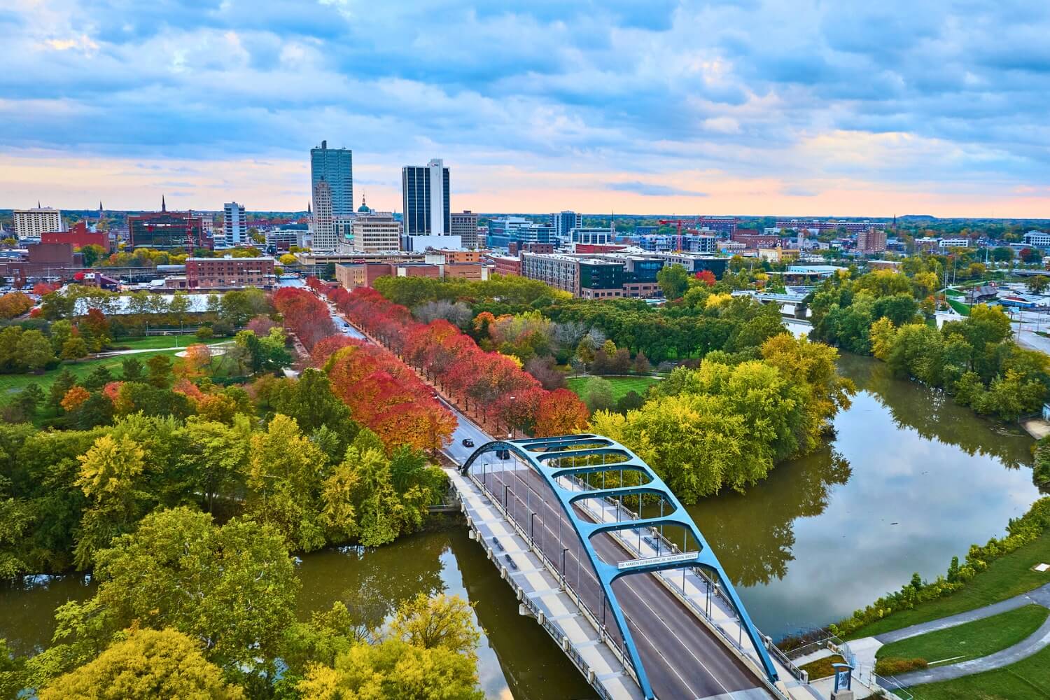 Aerial view of a cityscape during autumn, showing a blue arched bridge crossing a calm, tree-lined river. The trees display vibrant fall colors, especially along a straight path lined with red and orange foliage.