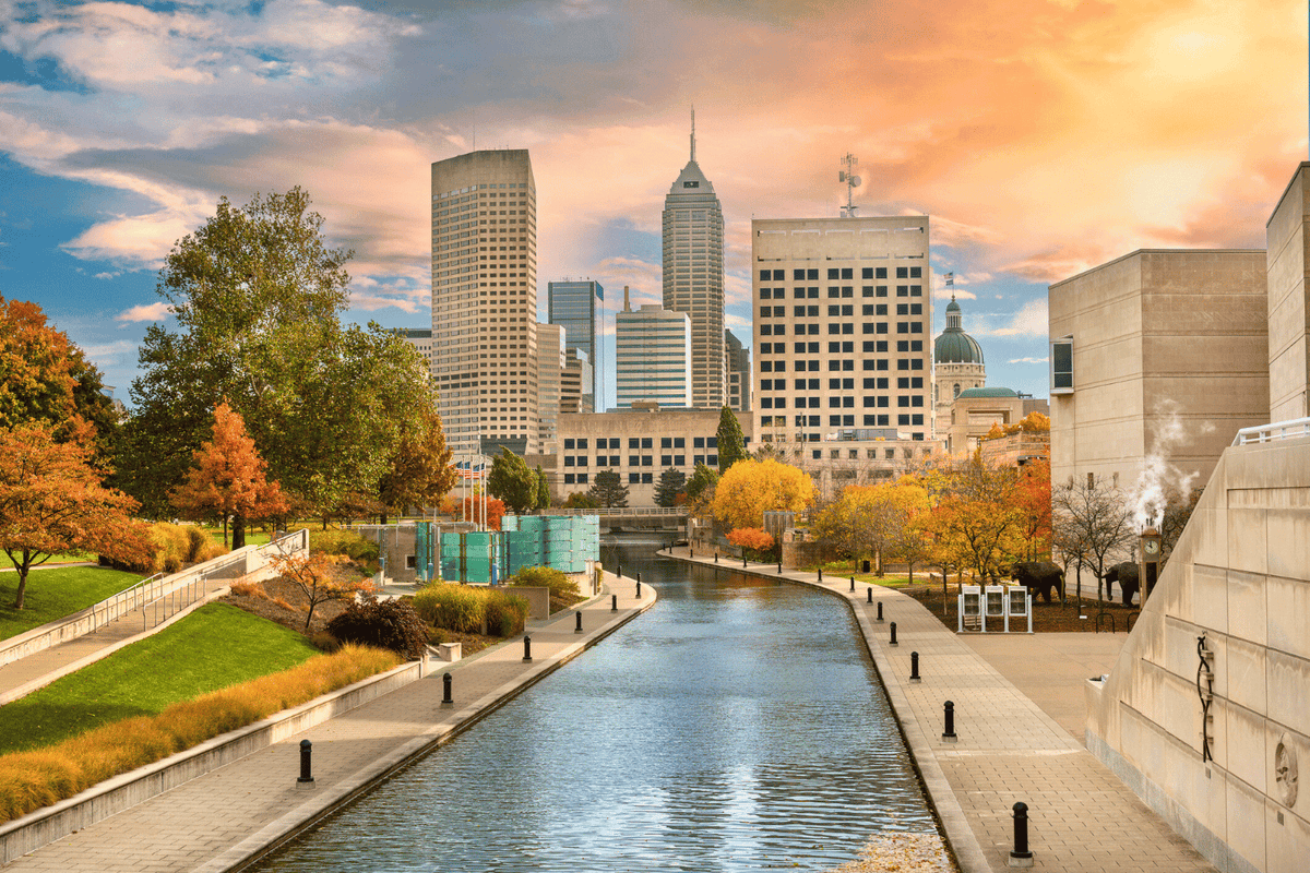 A scenic view of the Indianapolis Canal Walk during autumn, with the canal running through the center, flanked by walkways and vibrant fall foliage. In the background, the Indianapolis skyline rises against a dramatic orange and blue sunset sky.