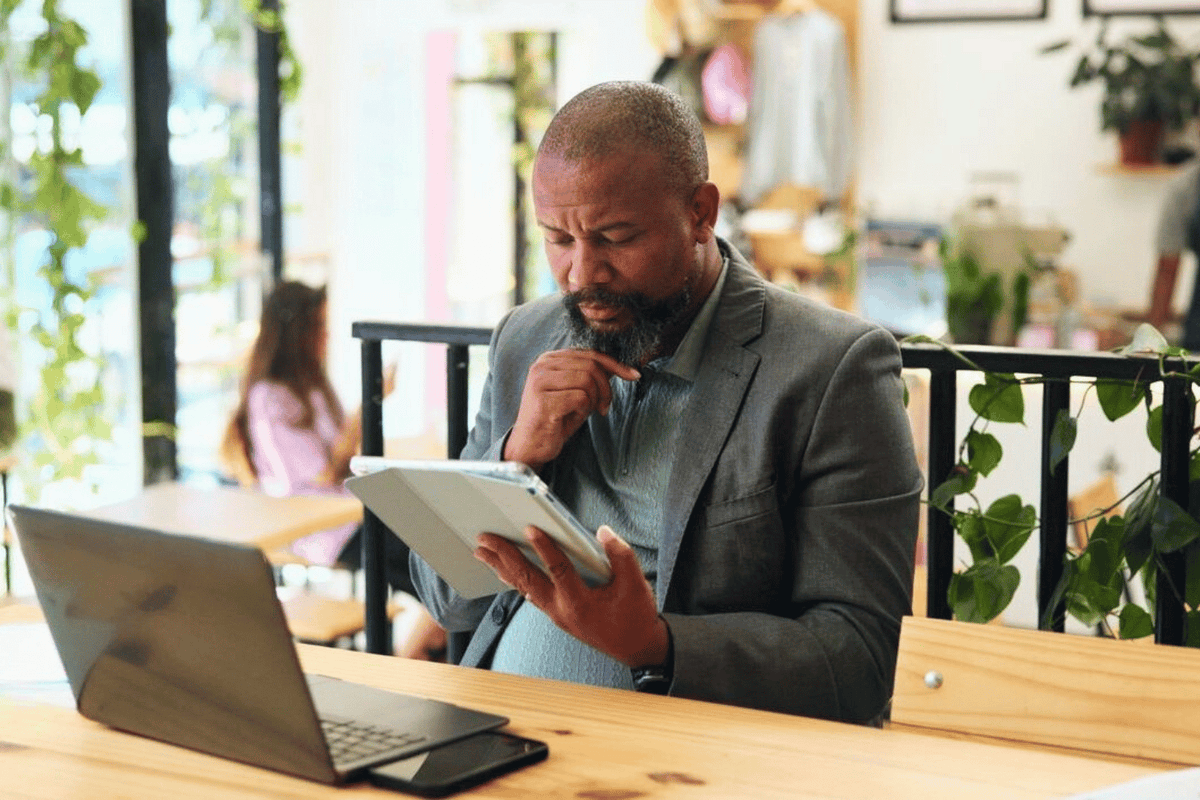 A man in a grey suit sits at a wooden table in a café, looking thoughtfully at a tablet in one hand while resting his other hand on his chin.