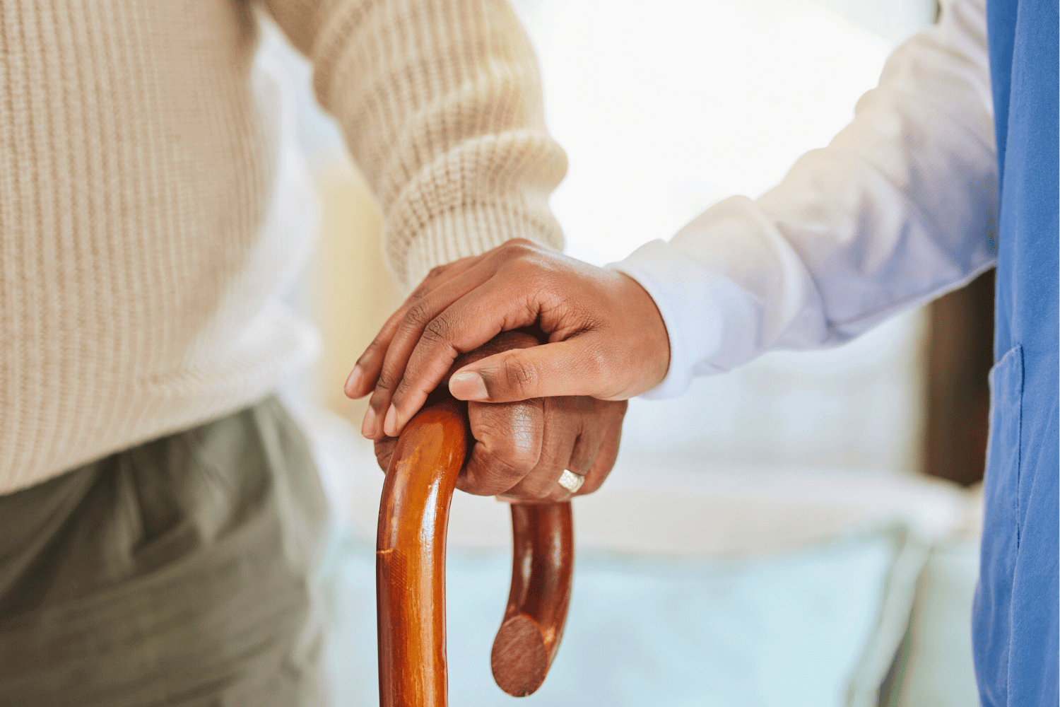 A close-up of a caregiver’s hand gently resting on top of an elderly person’s hand, which is gripping a wooden cane. The caregiver wears a blue uniform, and the elderly person wears a beige sweater.