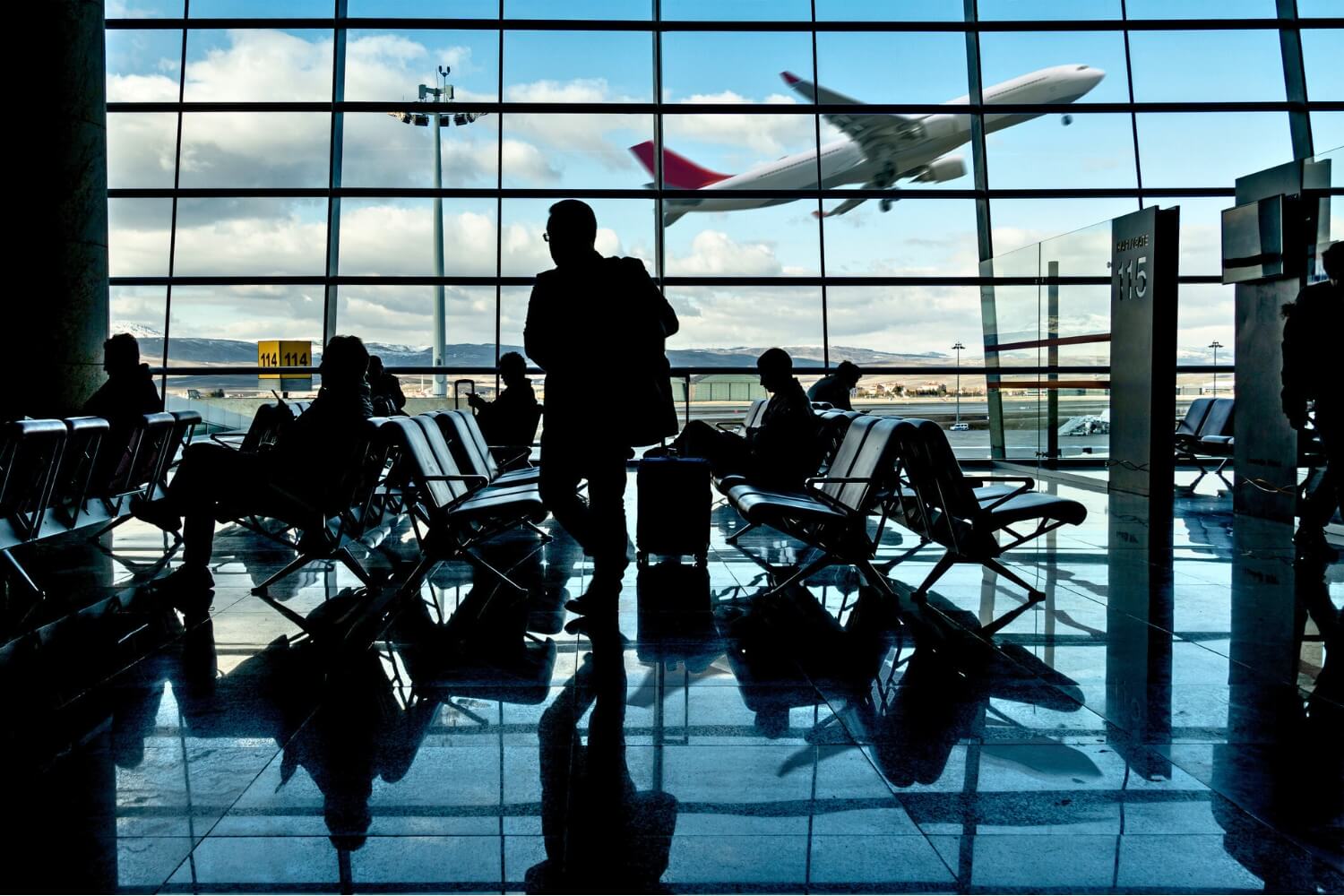 Passengers sit and stand with luggage in an airport departure lounge, silhouetted against large windows showing a plane taking off and a partly cloudy sky.