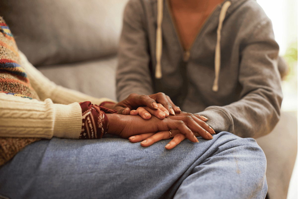 Close-up of two people sitting on a couch, one placing their hands gently over the other’s in a comforting gesture. Only their torsos and hands are visible; one person wears a gray hoodie and the other wears a colorful knit sweater 