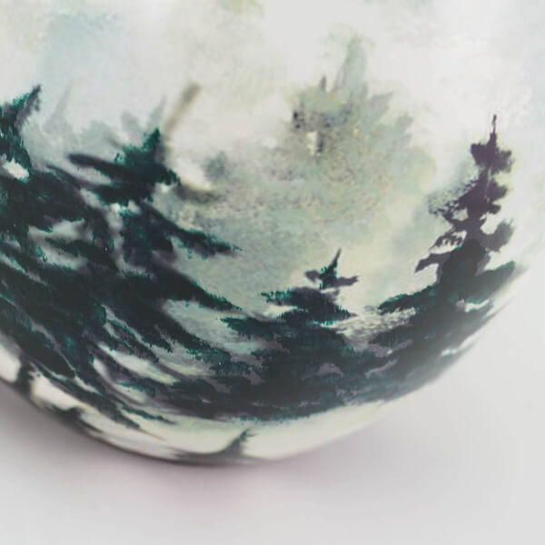 Close up of ceramic memorial cremation urn with nature-themed design of trees and water on a white background