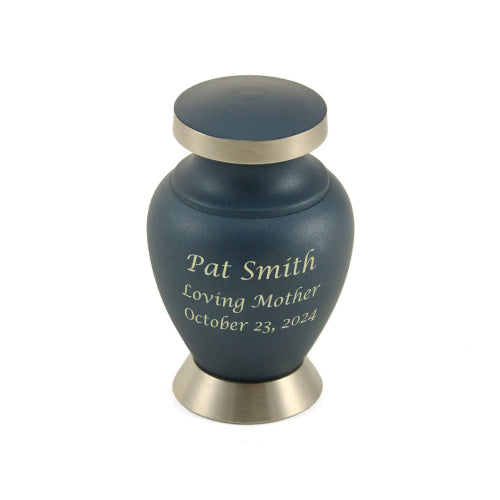 The Aria Ascending Keepsake Urn, available at Magnolia Cremations, is a small, navy blue urn with brushed silver accents and an engraved inscription. The urn has a smooth matte finish, a rounded shape, and a slightly flared base. Its silver-trimmed lid complements the minimalist and classic design. Engraving is shown as an available option.