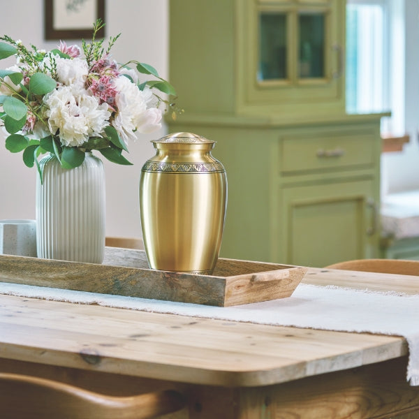 Elegant brass cremation urn displayed on a wooden tray with a floral arrangement in a home setting.