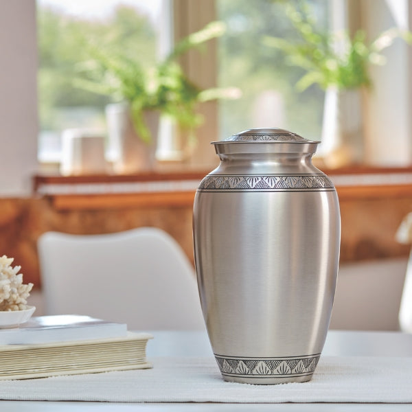 Elegant silver Classic Athena urn displayed on a table with natural light and greenery in the background.