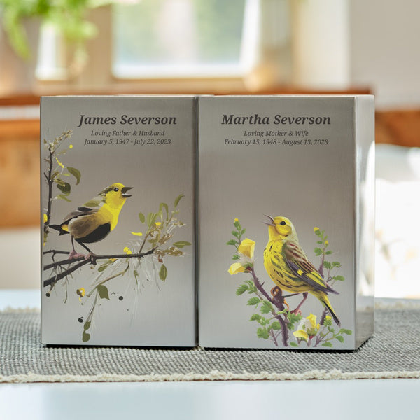 Life Canvas Birds Pewter Companion Urns displayed side by side. These rectangular stainless-steel cremation urns feature a brushed pewter finish with artistic illustrations of songbirds perched on tree branches. Designed for couples or loved ones to be placed together. Engraving is available as a personalization option. Secure bottom panel closure with four screws. Each urn has a 200 cubic inch capacity.