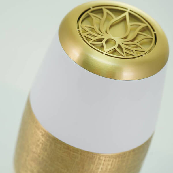 Close-up view of the top of a scattering urn, highlighting its gold-colored lid with an intricate lotus flower cutout design. The body of the urn is white with a textured gold band beneath the lid.