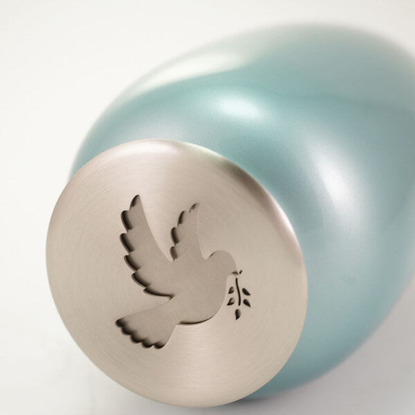 Close-up of a light blue scattering urn lying on its side, highlighting the silver-toned lid with a cutout design of a dove carrying an olive branch.