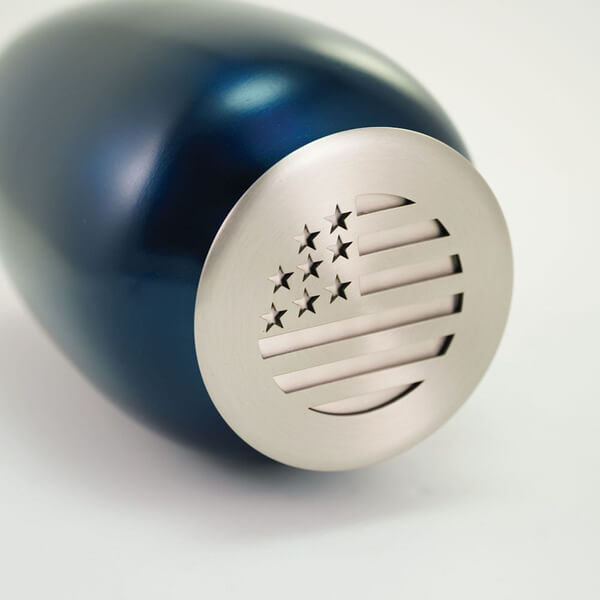 Close-up view of a dark blue scattering urn lying on its side, showcasing the silver-toned lid. The lid features a stylized cutout design of the American flag, with five stars and five horizontal stripes arranged in a circular pattern.
