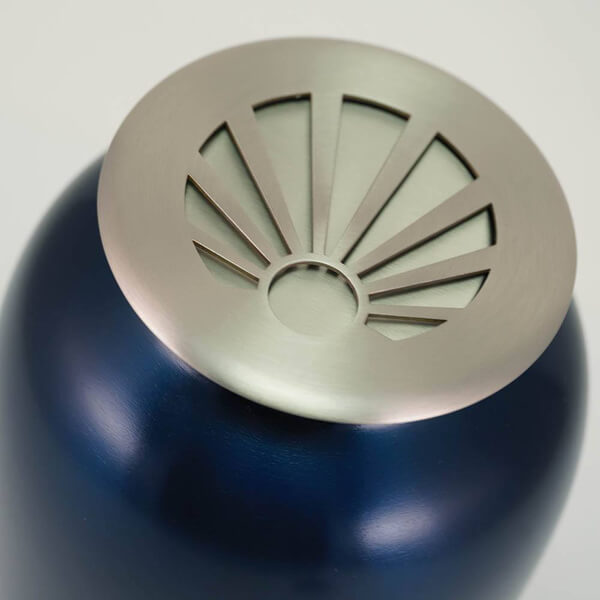 Close-up view of the lid on a dark blue scattering urn. The lid is metallic with a brushed finish and features a sunray design, revealing a lighter inner layer beneath.