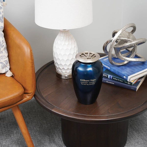 A dark blue scattering urn with a brushed metal lid sits on a round wooden side table. The urn is engraved. The table also holds a white textured lamp, a decorative metallic sculpture, and a stack of books. A brown leather chair is partially visible beside the table.