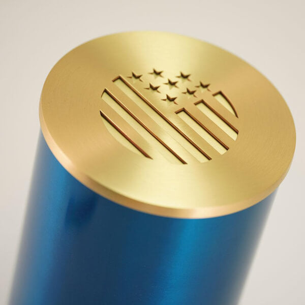 Close-up view of the lid of a cylindrical scattering urn. The lid is metallic gold with a precision-cut design resembling a stylized American flag, featuring stars and vertical stripes within a circular shape. The body of the urn is metallic blue.