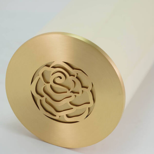 Close-up view of the lid of a scattering urn, showing a brushed gold finish with an intricately cut rose design in the center. The urn itself is a soft cream color, partially visible in the background.