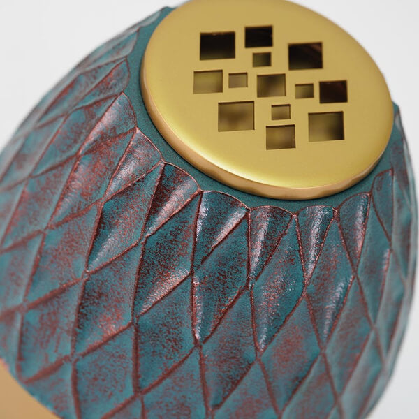 Close-up view of a decorative scattering urn with a textured, diamond-patterned surface in a distressed teal and reddish-brown finish. The urn has a gold-colored lid featuring a modern square and rectangular cutout design.