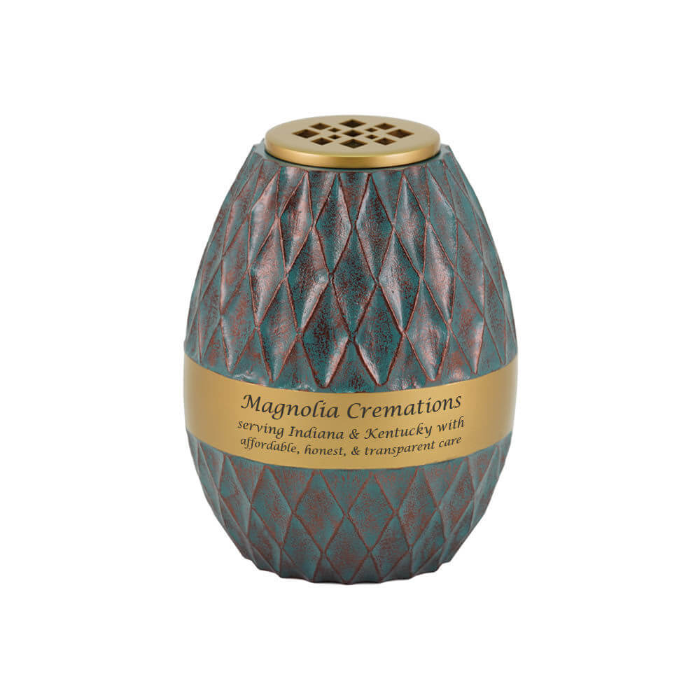Decorative scattering urn with a textured, diamond-patterned surface in teal and reddish tones, featuring a wide gold band around the middle. The band is engraved with the text: "Magnolia Cremations – serving Indiana & Kentucky with affordable, honest, & transparent care." The urn has a gold lid with a geometric cutout design.