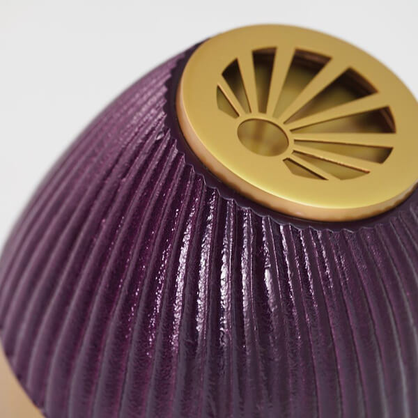 Close-up view of the top portion of a purple, ribbed scattering urn with a gold lid. The lid features a decorative sunburst cutout pattern, highlighting the texture and detail of both the urn and the lid.