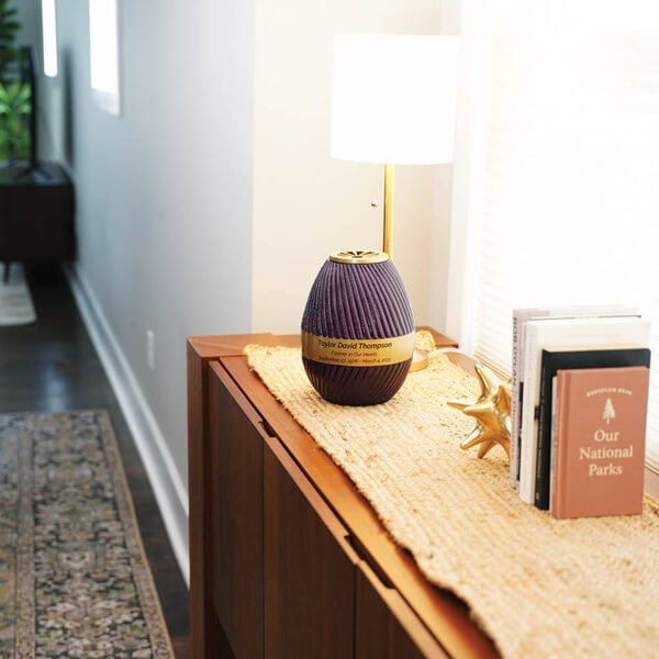 A purple, ribbed scattering urn with a gold band and nameplate sits on a woven table runner atop a wooden console table. The urn is displayed beneath a modern gold and white table lamp, next to a stack of books and a decorative gold starburst sculpture in a well-lit hallway or living space.