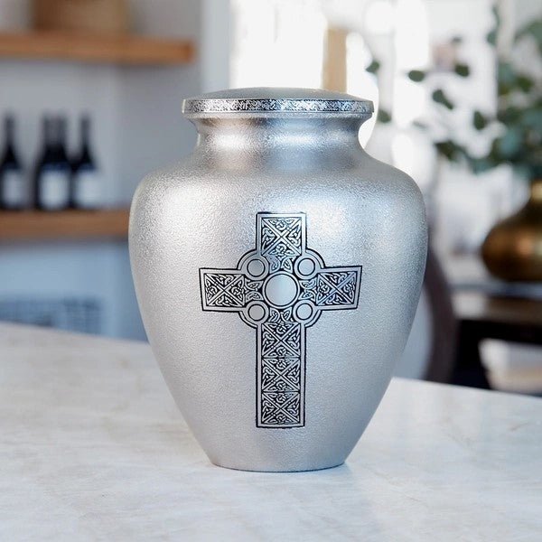 Celtic Cross Full-Size Cremation Urn displayed on a marble countertop in a home setting. This brass urn features a textured silver finish with an engraved Celtic Cross, symbolizing faith and eternity. The lid is decorated with an intricate Celtic pattern. 200 cubic inch capacity.