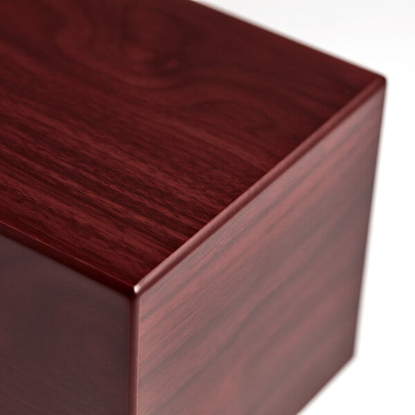 Close-up of the corner of a polished wooden cremation urn with a rich cherry-brown finish, showing detailed wood grain and smooth, glossy surfaces.