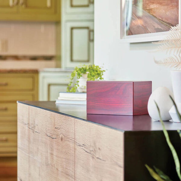 A polished wooden cremation urn with a reddish finish sits on a modern wood-and-black sideboard in a home interior. The background shows a kitchen with green cabinetry, a plant, and framed artwork on the wall above the urn