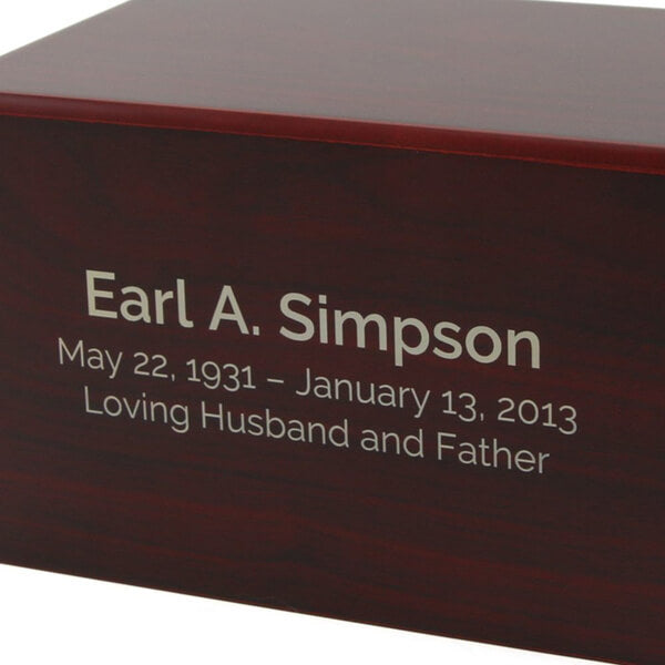 Close-up of a dark reddish-brown wooden cremation urn with an engraved inscription that reads: "Earl A. Simpson, May 22, 1931 – January 13, 2013, Loving Husband and Father."