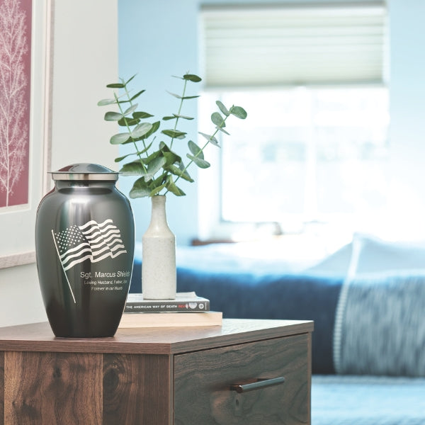 Classic Flag Full-Size Cremation Urn displayed on a wooden nightstand in a softly lit bedroom. This black stainless-steel urn features a silver engraving of the American flag, representing patriotism and honor. Custom engraving is available for personalization. 200 cubic inch capacity.