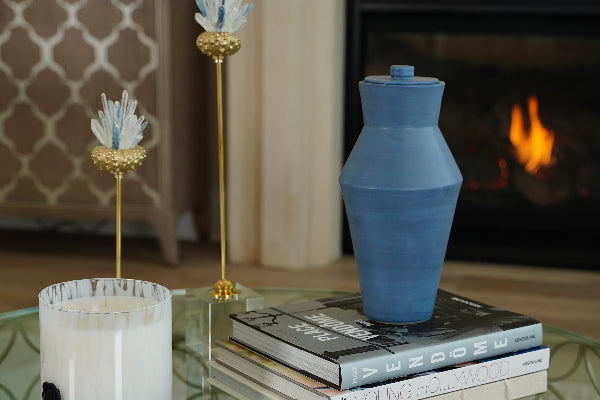 Deco Pure Sky Full-Size Cremation Urn by Stoneware & Co. displayed on a glass coffee table in a refined living room. The handcrafted ceramic urn in calming sky blue features an Art Deco-inspired design with clean lines. A warm fireplace and elegant decor create a peaceful ambiance. Suitable for individuals weighing up to 210 lbs.