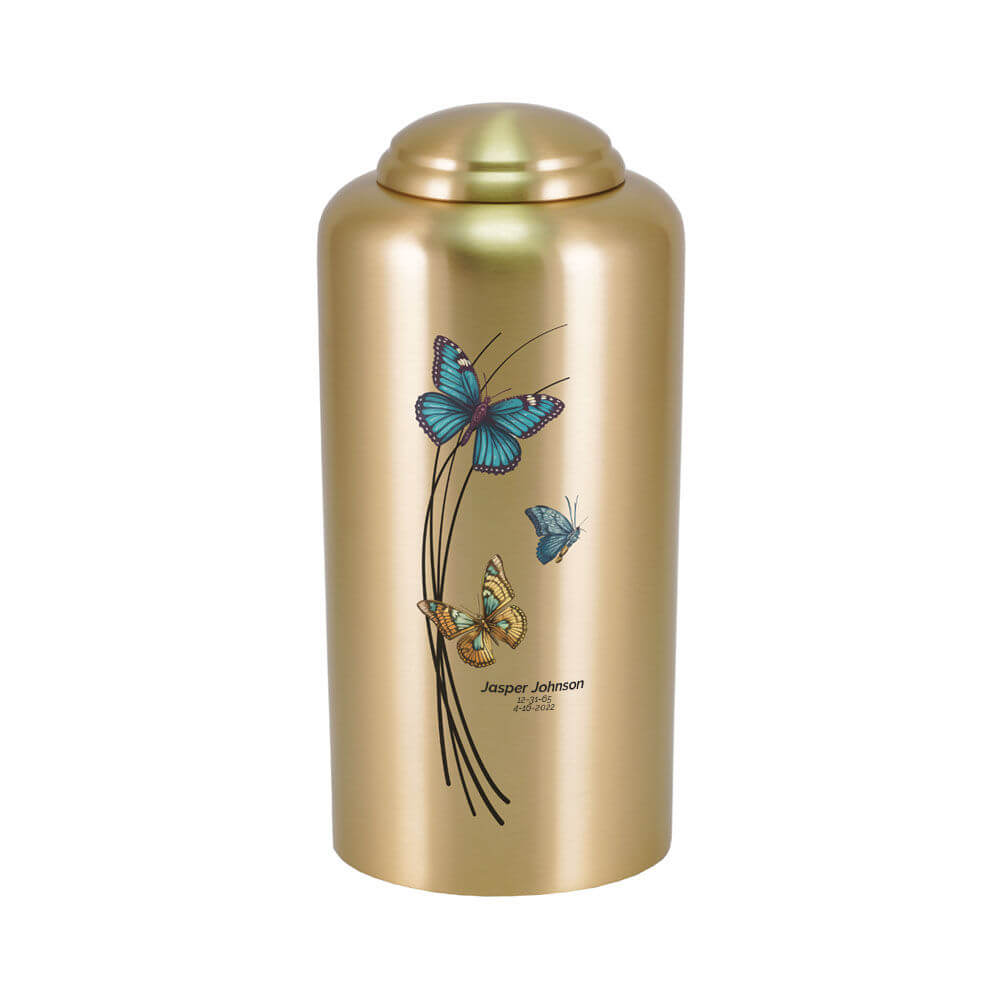 Gold-colored cremation urn with a domed lid, decorated with three colorful butterflies—blue, orange, and teal—flying around black curved lines. The urn also features optional engraving with a name and dates below the design.