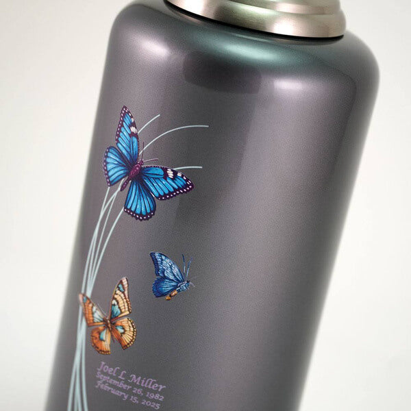 Close-up of a dark gray cremation urn with a metallic finish, showing three colorful butterflies—blue, teal, and orange—flying around pale green curved lines. Below the design, there is engraved text with a name and dates.