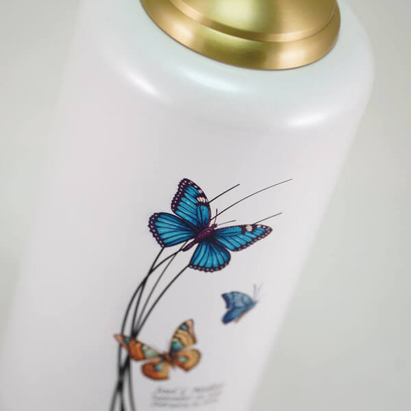 Close-up of a white cremation urn with a gold domed lid, showing three colorful butterflies—blue, teal, and orange—flying among black curved lines. Below the design, faint engraved text with a name and dates is visible.