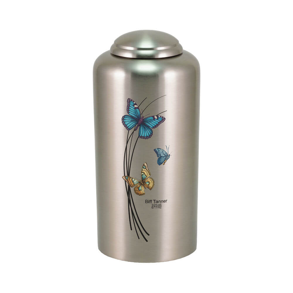 Silver-colored cremation urn with a domed lid, featuring a design of three butterflies—blue, teal, and orange—fluttering around black curved lines. The urn includes optional engraving below the design, displaying a name and dates.