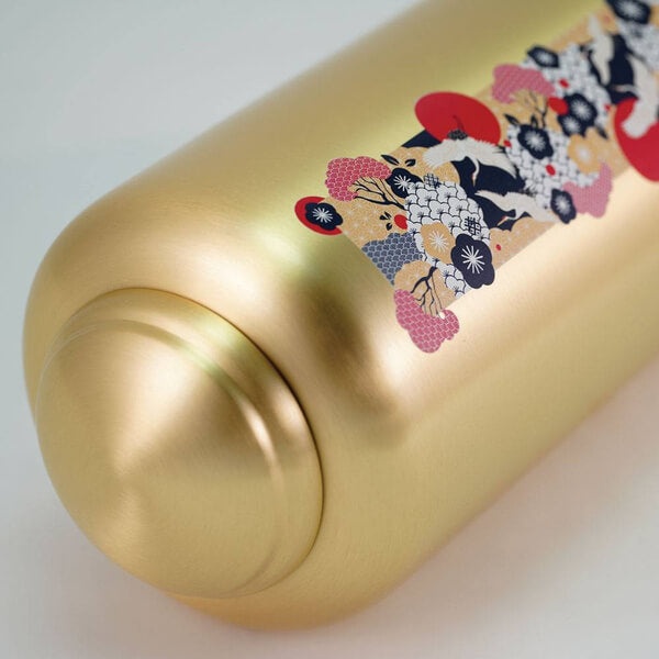 Close-up view of a gold-colored cremation urn lying on its side, highlighting a decorative vertical panel. The design features stylized cranes in black and white surrounded by red suns, floral patterns, and fan motifs in red, black, and gold tones, inspired by traditional Japanese art.