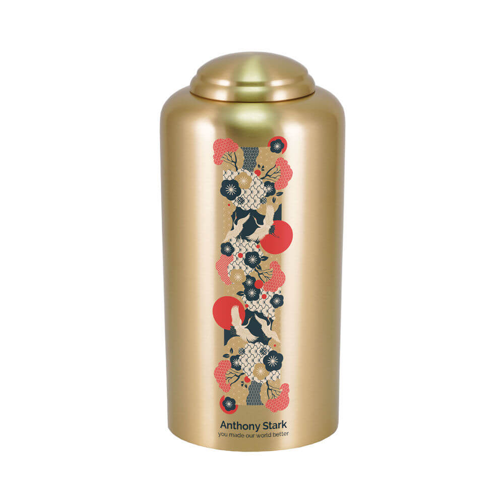 Gold-colored cremation urn with a domed lid, featuring a vertical decorative panel with a Japanese-inspired crane design. The pattern includes black and white cranes, red suns, and floral motifs in red, black, and gold. Below the design, there is optional engraved text displaying a name and personal message.