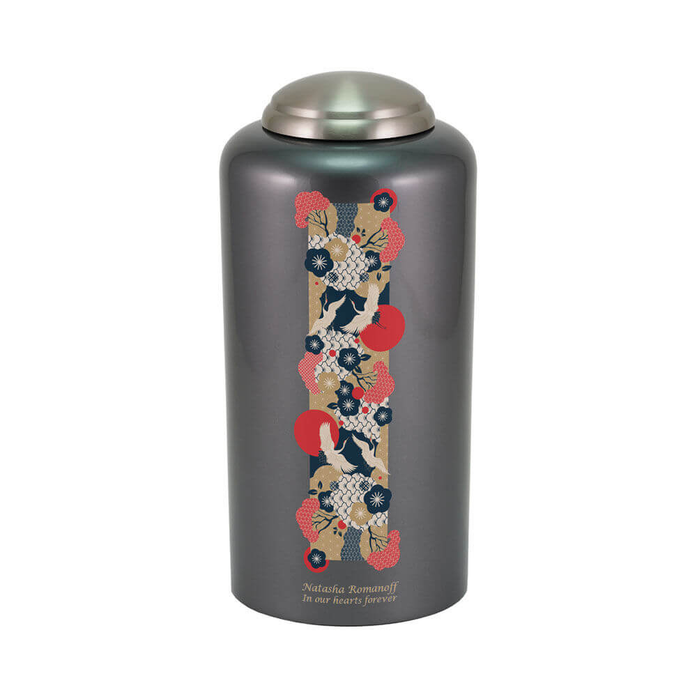 Dark gray cremation urn with a metallic lid, featuring a vertical decorative strip with an intricate Japanese-style design of cranes, red sun motifs, and floral patterns in red, navy blue, white, and beige. Near the bottom, engraved text is shown. 