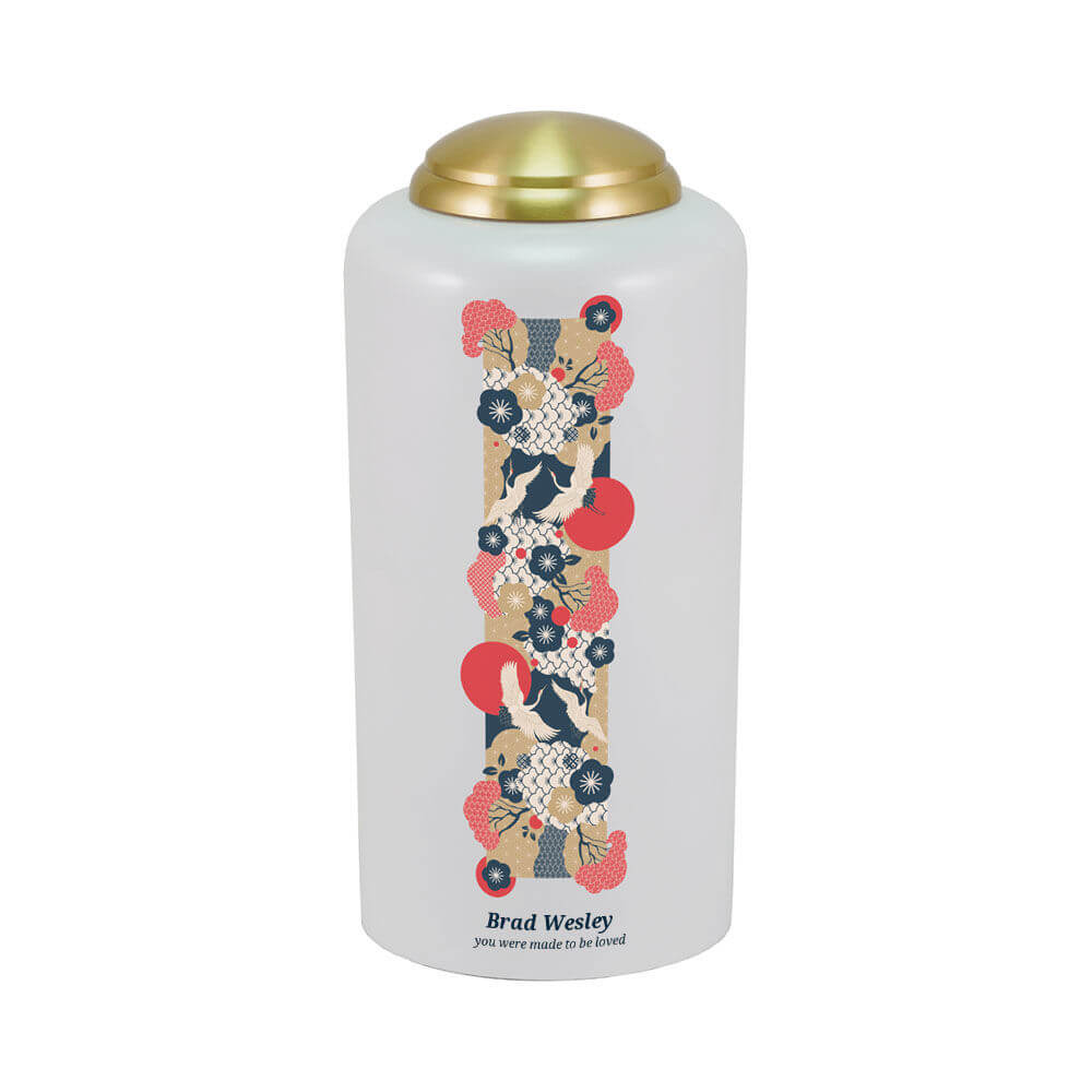 White cremation urn with a gold lid, featuring a vertical Japanese-style design of white cranes, red sun motifs, and floral patterns in red, navy blue, cream, and gold. Available optional engraving is shown.