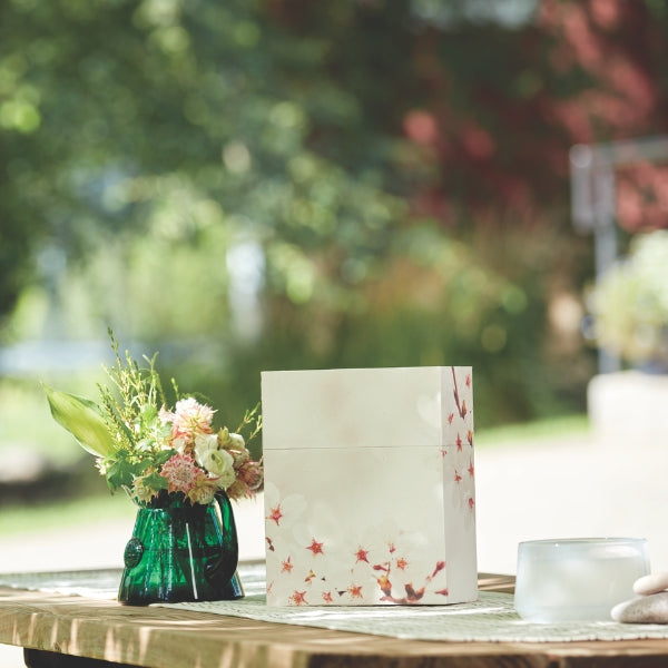 An eco-friendly memorial urn with a cherry blossom design, placed on an outdoor table next to a green glass vase with fresh flowers. The urn has a rectangular shape with a two-piece construction, designed for scattering or burial. Engraving is not available.
