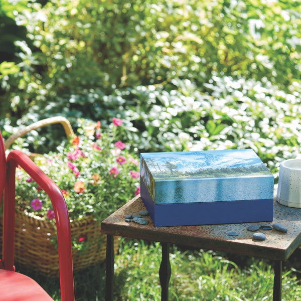 A rectangular eco-friendly memorial urn with a serene mountain and lake scene printed on the lid, placed on a rustic outdoor table. Small stones are arranged nearby, and a red chair and a basket of flowers sit in the background, surrounded by lush greenery.