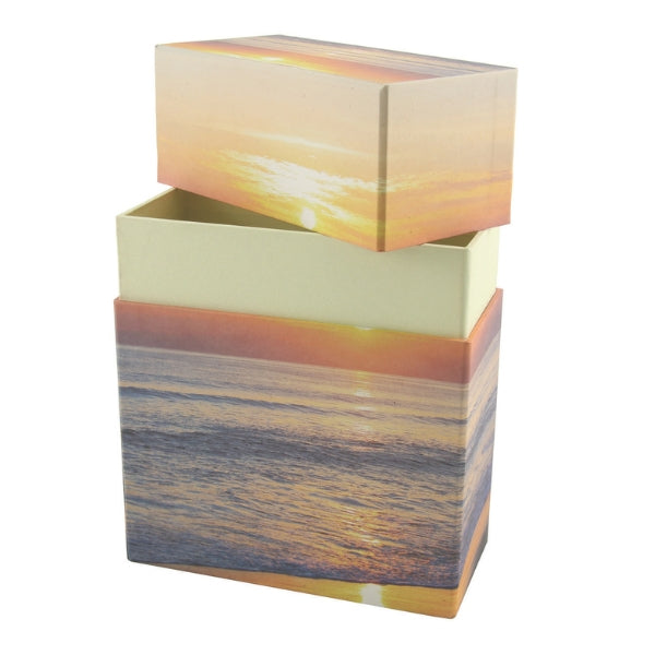 A biodegradable memorial urn featuring a peaceful beach and sunset design, shown with its lid removed to reveal a light-colored inner compartment. The urn is rectangular in shape and made of eco-friendly material, designed for scattering or burial.