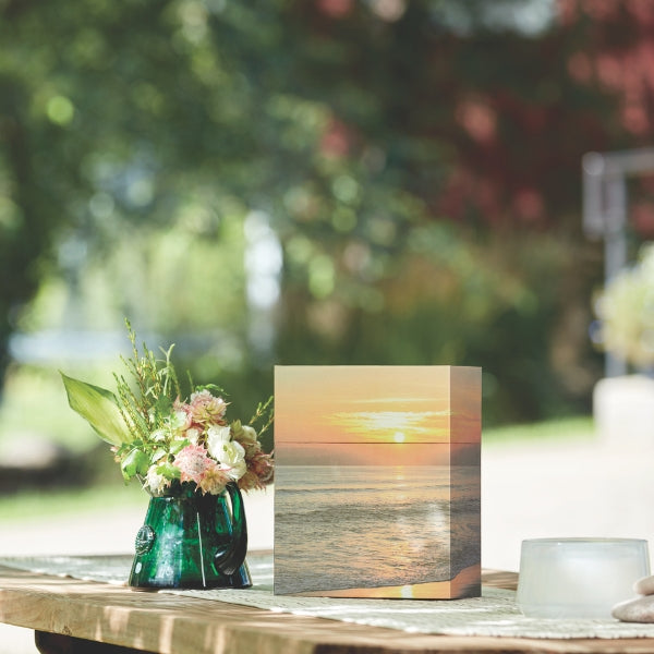 A biodegradable memorial urn with a printed image of a tranquil beach at sunset, placed on an outdoor table. The urn has a rectangular shape and is positioned beside a green glass vase with fresh flowers. Sunlight filters through the trees in the background, creating a peaceful setting.