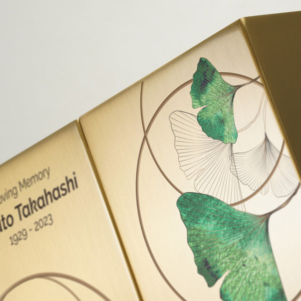A close-up of two golden memorial urns placed side by side, featuring a delicate ginkgo leaf design. The brushed metallic surface is adorned with green leaves and fine line illustrations, creating an elegant and artistic tribute.