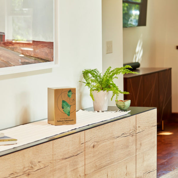 A golden memorial urn with a ginkgo leaf design is displayed on a modern wooden console table. The urn has a brushed metallic finish with green leaves and delicate circular linework. A potted fern and a small decorative bowl sit beside it, adding warmth to the serene setting.