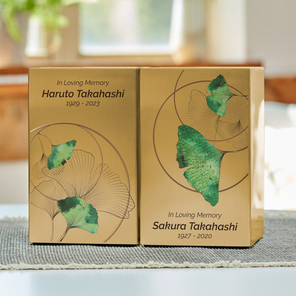 A pair of golden memorial urns placed side by side on a table, each engraved with a name and dates. The urns feature a ginkgo leaf design with green accents and fine line illustrations, symbolizing remembrance and longevity. Natural light filters through a window in the background, casting a warm ambiance.