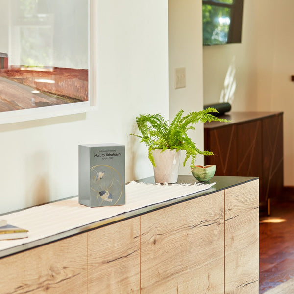 A silver-colored memorial urn with an elegant ginkgo leaf design sits on a modern wooden sideboard, accompanied by a green potted fern and a small ceramic bowl. The urn has a brushed stainless steel finish with gold detailing and textured blue and gold leaves. The space is bright and contemporary, with natural light filtering in from a nearby window.