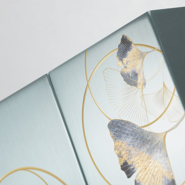 A close-up of two silver-colored companion urns featuring an intricate ginkgo leaf design in gold and blue tones. The metallic surface reflects light, highlighting the delicate pattern. Engraving is visible on one urn, with personalization available.