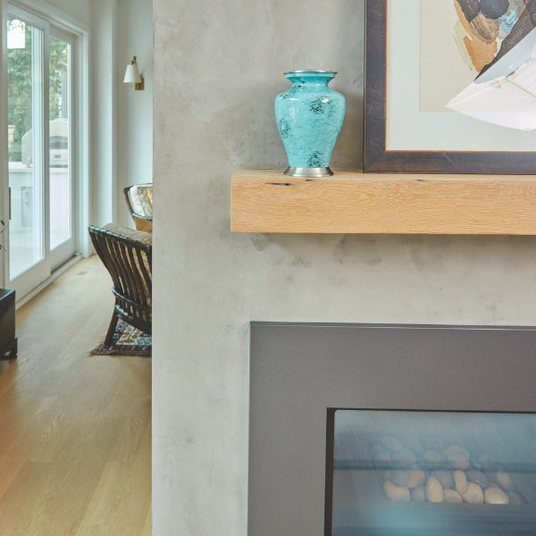 A small teal blue marble-patterned urn with silver-tone accents sits atop a wooden fireplace mantel. The urn has a classic, flared shape and a glossy finish. The modern living space features a concrete-style wall, a framed abstract artwork, and a black fireplace with decorative stones inside. Natural light streams in from large glass doors leading outside.