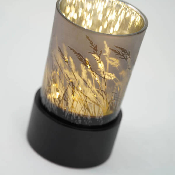 Close-up of a keepsake urn that also functions as a tealight candle holder. It features a frosted glass cup with a design of silhouetted grasses, glowing warmly from the candlelight inside, and sits on a round black base.