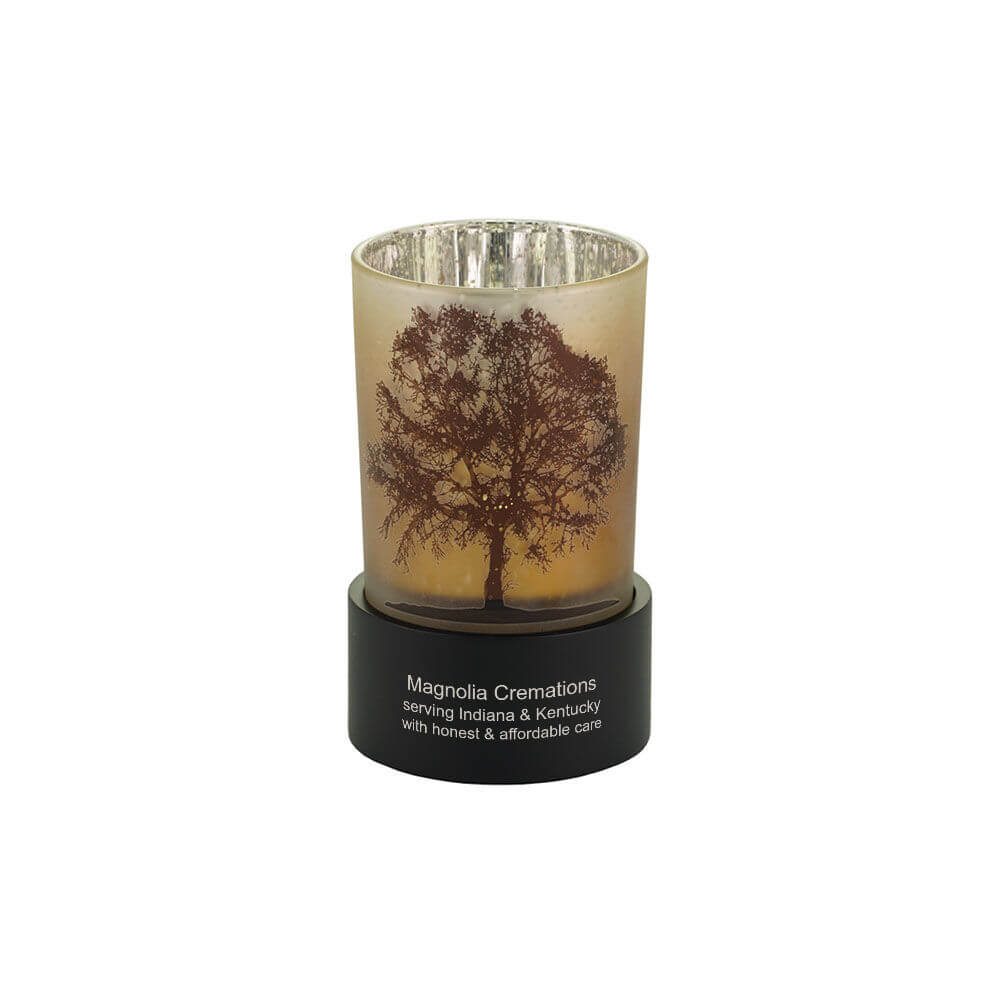 Keepsake urn that also functions as a tealight candle holder, featuring a frosted glass cup with a detailed tree silhouette and a black circular base. Optional engraving is shown.